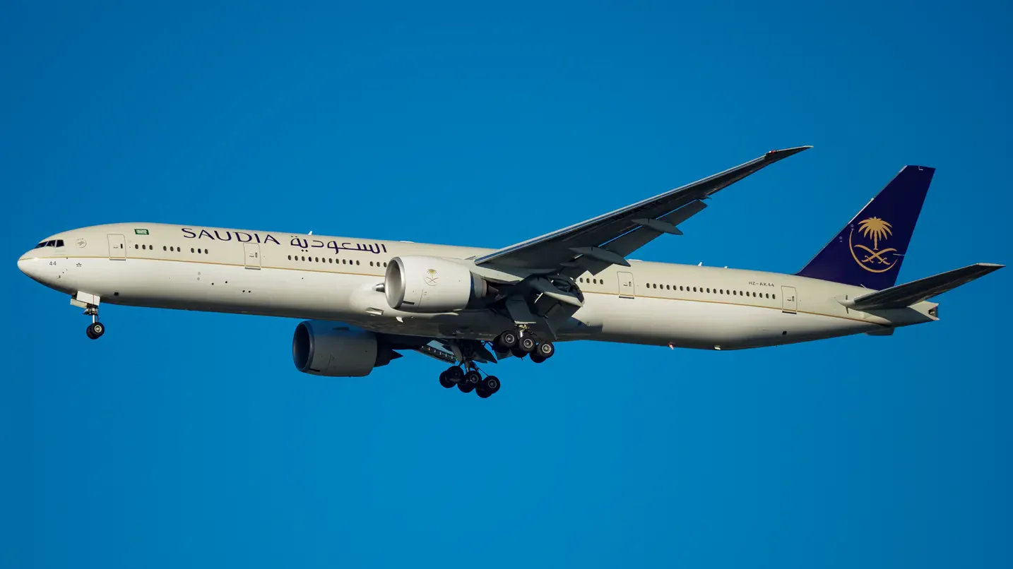 Saudia Boeing 777-300ER airplane in flight with landing gear down against a clear blue sky.