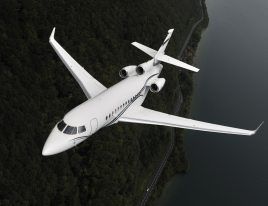 Dassault Falcon 7X private jet in flight over a river and forested terrain, viewed from above