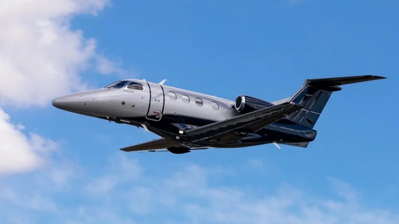 Embraer Phenom 100 private jet in flight against blue sky, side profile view showing silver fuselage and dark tail.