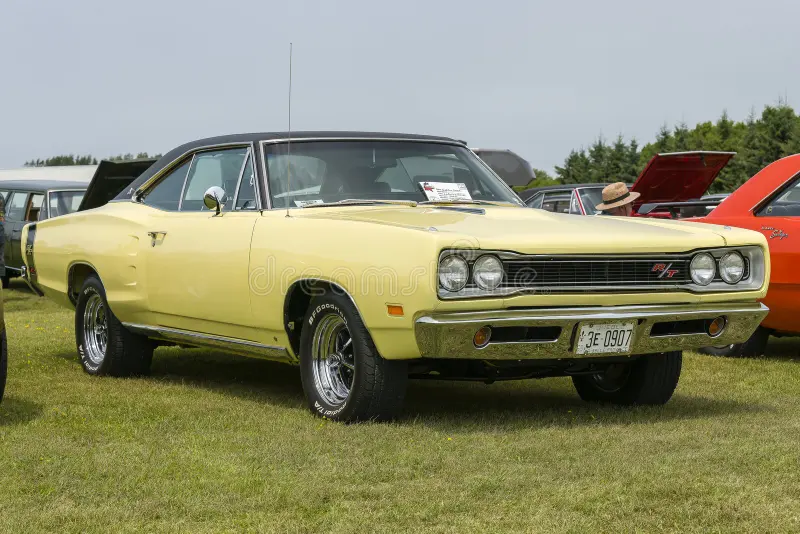 Classic muscle car with a yellow exterior and black vinyl roof, chrome detailing, and dual exhausts, parked on grass. Features iconic round headlights, prominent front grille, and a badge. The wheels are chromed with white-lettered tires. The license plate is visible on the front bumper. The car's length and design reflect late 1960s American automobile styling. Other vintage vehicles and people are visible in the background, indicating a car show setting. Classic muscle cars stock images, royal