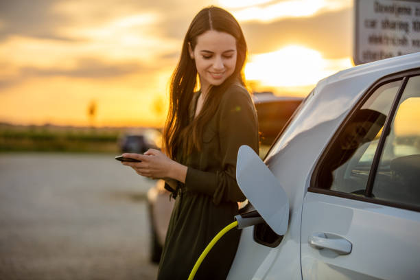 Woman waiting for electric car to charge in the parking lot at sunset Side view waist up shot of a young woman in a dress using her mobile phone while waiting for her white electric car to charge during sunset electric car exterior stock pictures, royalty-free photos & images
