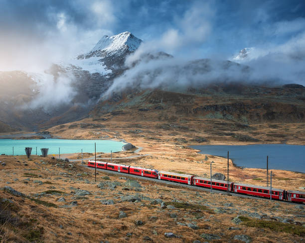 Aerial view of red train near alpine mountain lake and cloudy sky Aerial view of red Bernina Express train, curving railroad, turquoise lake in the Swiss Alps, rocky hills and dramatic cloudy sky at sunset in autumn. Alpine mountains. Top view of train in fall glacier express switzerland stock pictures, royalty-free photos & images