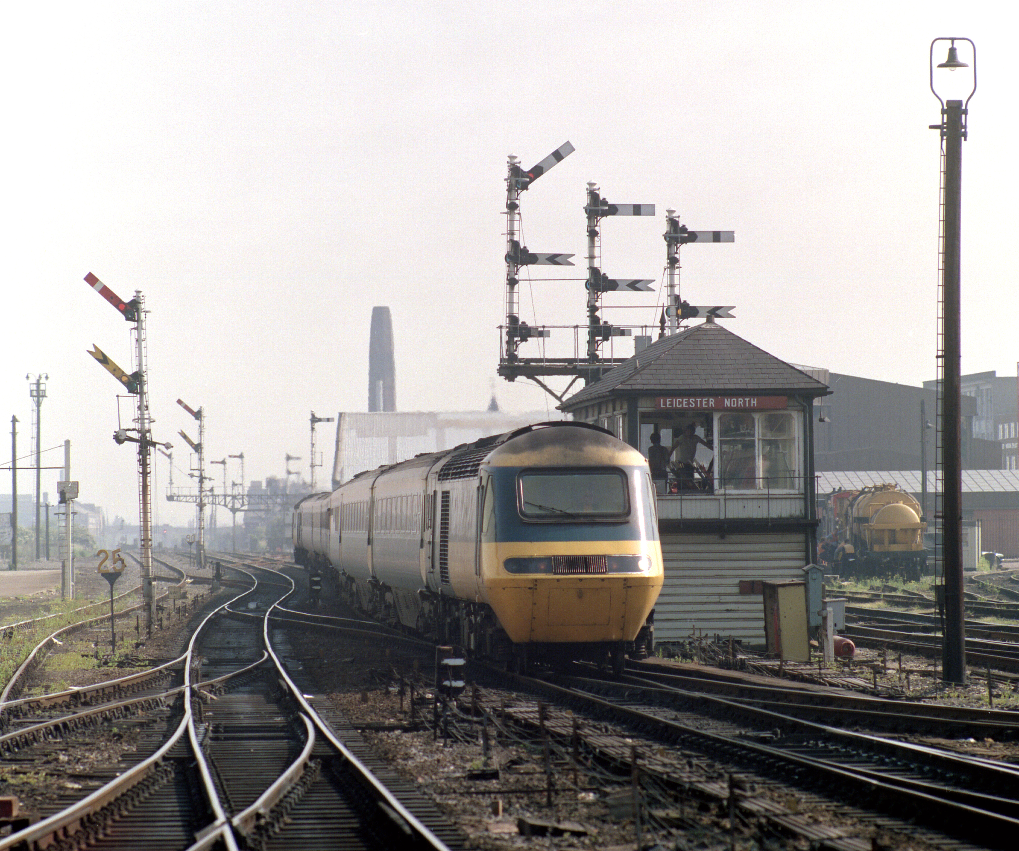 An InterCity 125 enters a station, taken almost head on. It is surrounded by semaphore signals behind it and the tracks are beginning to diverge. On its right side (i.e. the driver's left) there is an old-style signal box whose height is barely taller than the train.